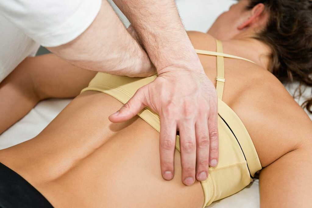 A person sitting on a couch, holding their lower back with one hand and massaging their thigh with the other.